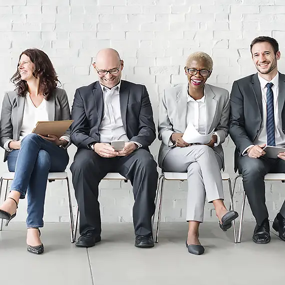 Four diverse professionals in suits sit, waiting, some smiling, holding papers/devices against a white brick wall.