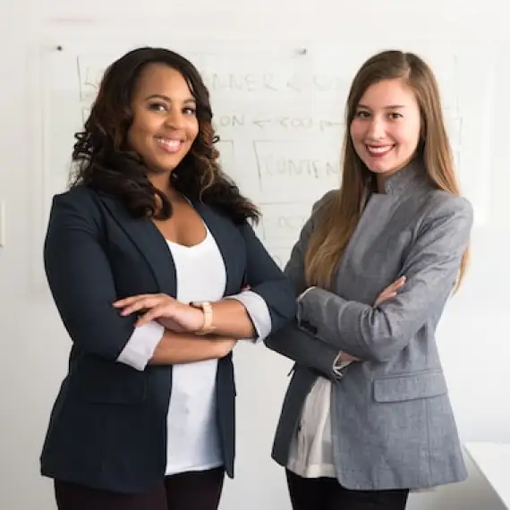 Two smiling women, likely in a professional setting. They are standing with their arms crossed, suggesting confidence and a collaborative dynamic. The background is a plain white wall or whiteboard.